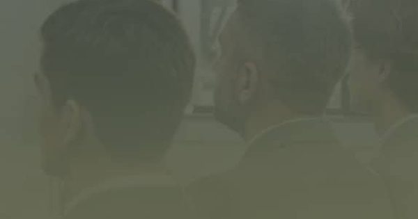 Three men in suits sit indoors, facing a whiteboard or screen, with a window and curtain visible in the background.