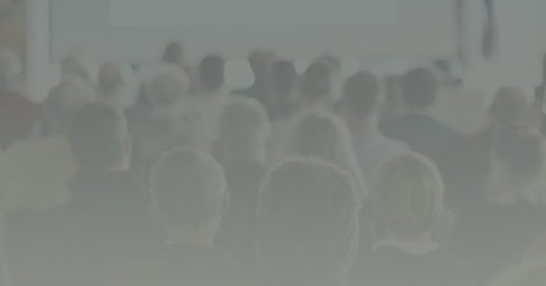 A group of people sit facing a large blank projection screen in a conference or seminar setting.