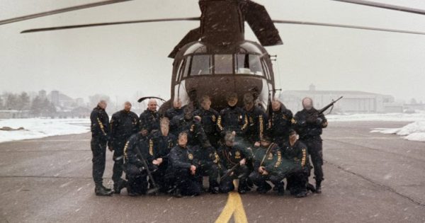 A group of uniformed individuals poses in front of a helicopter on a snowy tarmac, with snow falling and buildings in the background.