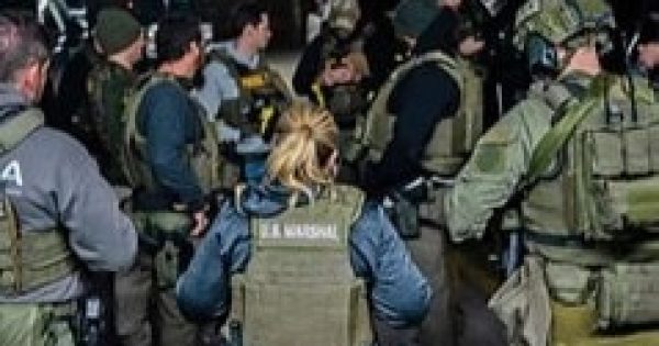 A group of law enforcement officers in tactical gear, including one with a visible "U.S. Marshal" vest, stand together during an operation.